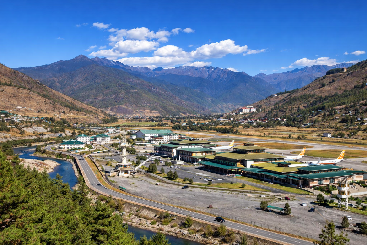 Panoramic view of Paro Valley in Bhutan, featuring Paro International Airport with traditional Bhutanese architecture, a winding river, surrounding roads, and dramatic Himalayan mountains under a clear blue sky.